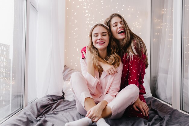Linda garota europeia em pijamas rosa, posando com um sorriso encantador no quarto. Retrato interno de mulheres fascinantes de pijama se abraçando e rindo.
