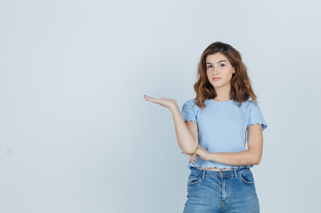 Linda garota fazendo o gesto de boas-vindo em t-shirt, jeans e olhando confiante, vista frontal.