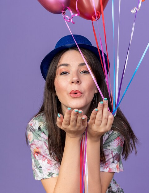 Linda garota feliz com chapéu de festa segurando balões, mostrando gesto de beijo isolado na parede azul