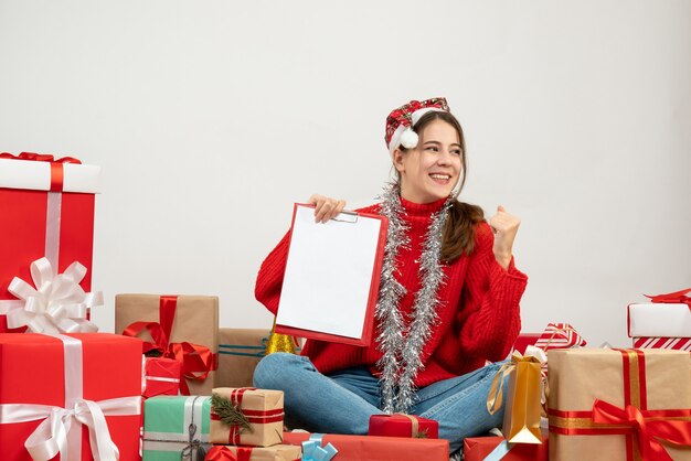 linda garota feliz com chapéu de Papai Noel segurando um documento sentado em volta de presentes em branco