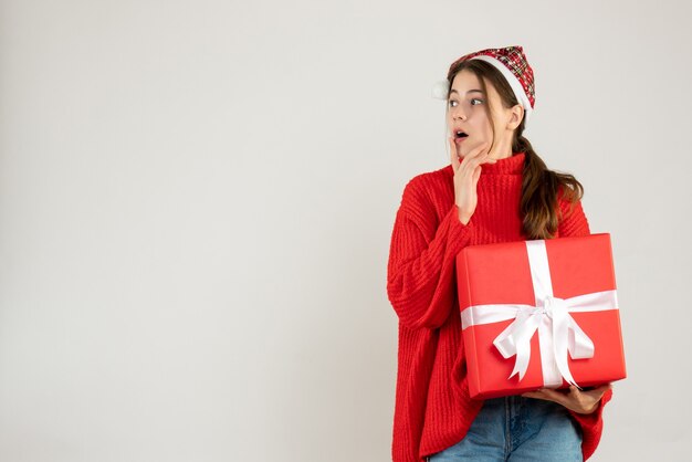 linda garota feliz com chapéu de Papai Noel segurando um presente, colocando a mão no queixo em pé no branco