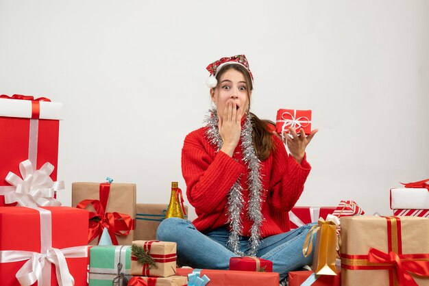 Linda garota festeira com chapéu de Papai Noel segurando um presente, colocando a mão na boca, sentada em volta dos presentes