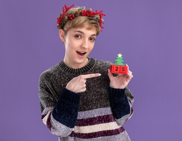 Linda garota impressionada com uma coroa de flores na cabeça e segurando um brinquedo da árvore de natal com a data apontando para ele olhando para a câmera isolada no fundo roxo