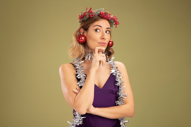 Linda garota impressionada com vestido roxo e grinalda com guirlanda no pescoço segurando bolas de natal nas orelhas agarrando o queixo isolado no fundo verde oliva