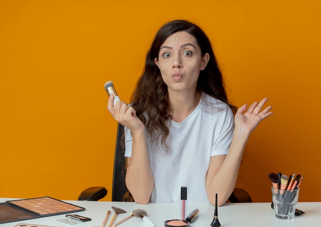 Linda garota impressionada sentada à mesa de maquiagem com ferramentas de maquiagem, mantendo as mãos no ar e segurando o pincel de base com creme de base colocado no rosto isolado em um fundo laranja
