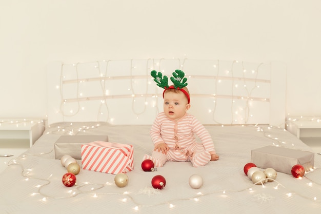 Linda garota infantil sentada na cama de decorações de Natal e vestindo o dorminhoco do bebê listrado de manga comprida e chifres de veado festivos, olhando para a câmera, comemorando o ano novo.