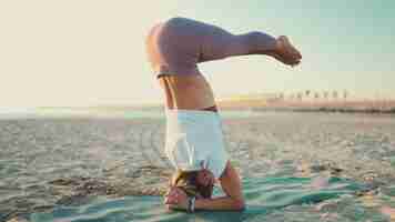 Foto grátis linda garota iogue forte fazendo parada de cabeça enquanto pratica poses de ioga à beira-mar mulher bonita, mantendo o equilíbrio durante a prática de ioga matinal na praia