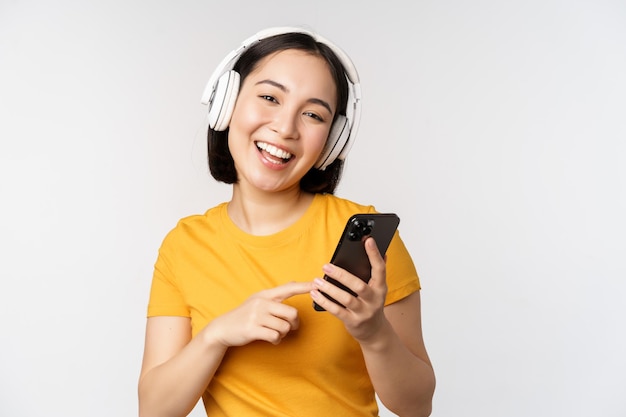 Linda garota japonesa em fones de ouvido olhando para o celular e sorrindo usando o aplicativo de música no smartphone em pé contra o fundo branco