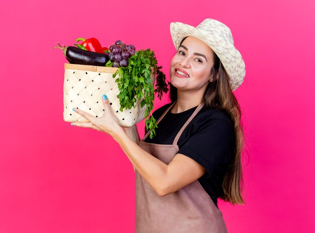Linda garota jardineira sorridente de uniforme usando chapéu de jardinagem levantando cesta de vegetais