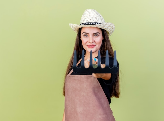 Foto grátis linda garota jardineira sorridente de uniforme usando chapéu de jardinagem segurando um ancinho para a câmera isolada em fundo verde oliva