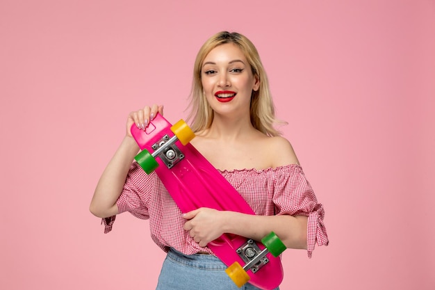 Linda garota loira adorável com batom vermelho na blusa rosa segurando o skate rosa alegremente