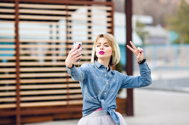 Linda garota loira elegante está se divertindo e faz selfie com um beijo na cara e com os dedos do sinal de vitória com bloqueios de madeira marrom listrada