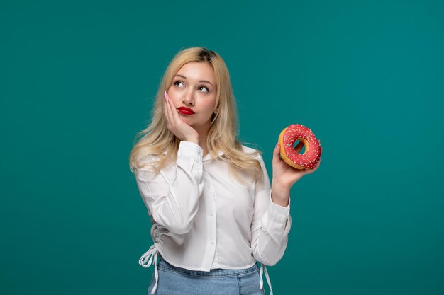 Linda garota loira linda jovem em uma camisa branca pura hesitando em comer um donut