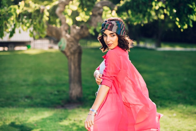 Linda garota morena com bandana andando no parque de verão. ela usa roupas brancas, sua longa camisa rosa está voando com o vento. vista de trás.