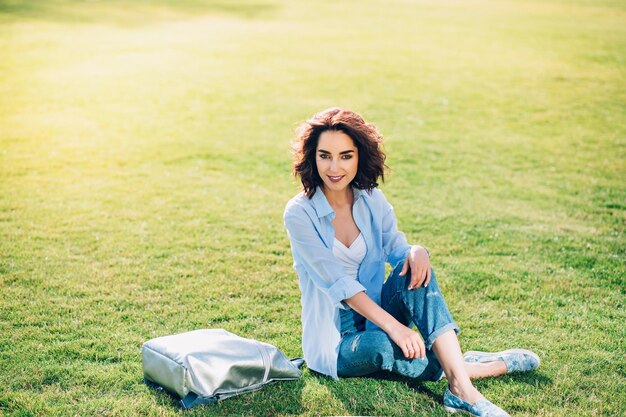 Linda garota morena com cabelo curto está posando na grama do parque. Ela usa camiseta branca, camiseta e calça jeans, sapatos. Vista de cima.