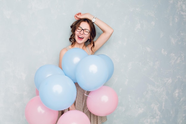 Linda garota morena de pé em um estúdio, sorrindo amplamente e segurando balões azuis e rosa. ela usa vestido claro pontilhado e óculos pretos, e tem cabelo trançado. ela está se divertindo.