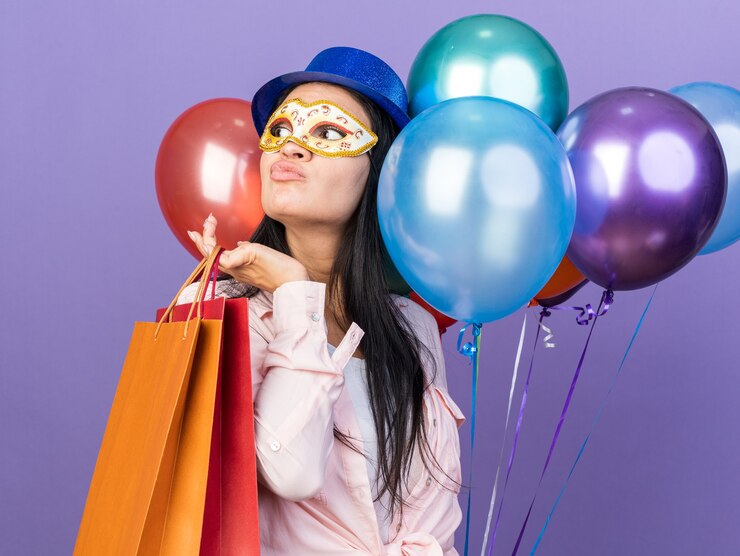 Foto grátis linda garota olhando para o lado com um chapéu de festa e máscara de baile de máscaras segurando balões com uma sacola de presente