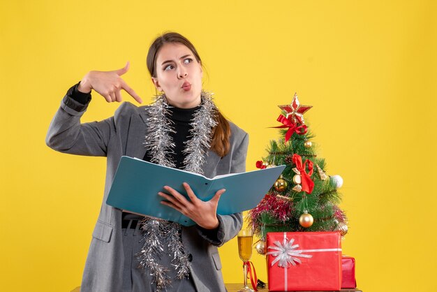 Linda garota pensativa de frente segurando documentos em pé perto da árvore de natal e coquetel de presentes