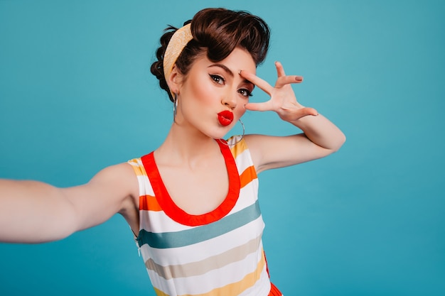 Linda garota pin-up se divertindo em fundo azul. Mulher bonita em vestido listrado, tomando selfie com o símbolo da paz.