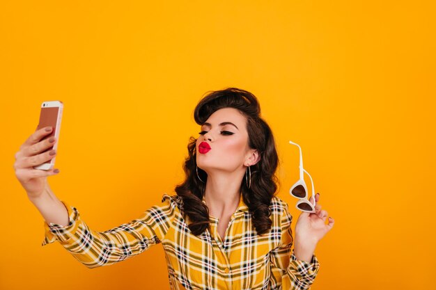 Linda garota posando com smartphone em fundo amarelo Pinup mulher tomando selfie com expressão de rosto beijando