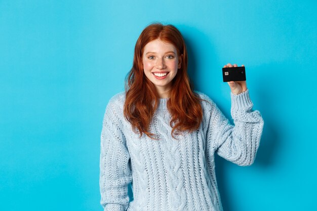 Linda garota ruiva com suéter mostrando cartão de crédito, sorrindo para a câmera, em pé sobre um fundo azul