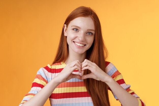 Foto grátis linda garota ruiva sorridente carismática de aparência amigável expressar afeto, amor e amizade, sorrindo, mostrar o sinal de coração no peito mostrando simpatia, confessar sentimentos românticos, fundo laranja de pé.