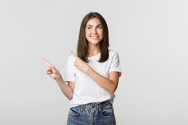 Foto grátis linda garota sonhadora sorrindo, apontando o dedo para o canto superior esquerdo e parecendo satisfeita com o logotipo