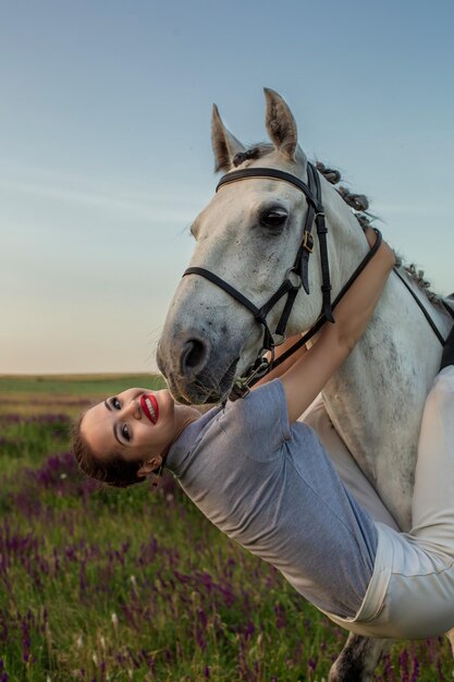 Linda garota sorri para seu cavalo vestindo competição uniforme: retrato ao ar livre no pôr do sol. Cuidando de animais, conceito de amor e amizade.