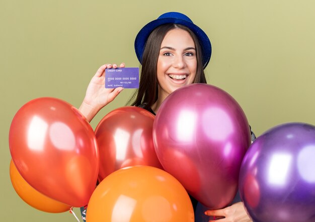 Linda garota sorridente com chapéu azul em pé atrás de balões segurando um cartão de crédito isolado na parede verde oliva