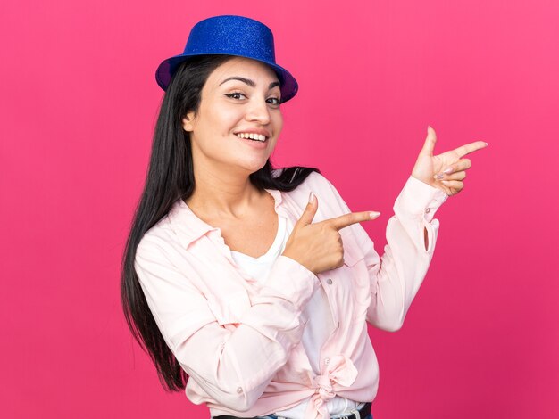 Linda garota sorridente com chapéu de festa apontando para o lado isolado na parede rosa