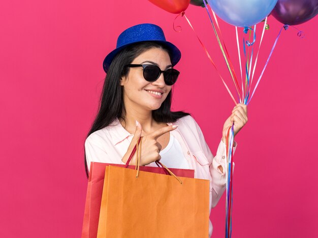 Foto grátis linda garota sorridente com chapéu de festa segurando balões e sacolas de presentes apontando ao lado