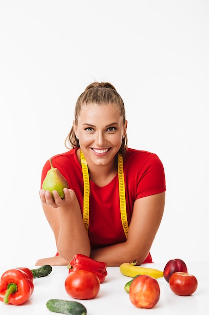 Linda garota sorridente com fita métrica no pescoço segurando pêra na mão enquanto alegremente olhando na câmera com frutas e legumes perto sobre fundo branco