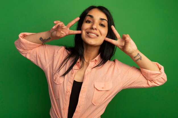 Linda garota sorridente com uma camiseta rosa mostrando um gesto de paz isolado na parede verde