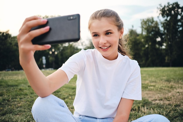Linda garota sorridente em camiseta branca feliz tirando foto no celular enquanto está sentado no gramado no parque da cidade