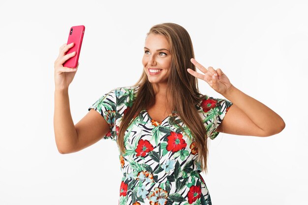 Linda garota sorridente em vestido colorido, mostrando o gesto de dois dedos enquanto alegremente tirando fotos no celular sobre fundo branco