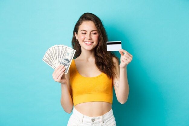 Linda garota sorridente mostrando dinheiro, mas olhando para o cartão de crédito de plástico com cara determinada e satisfeita, de pé sobre um fundo azul.