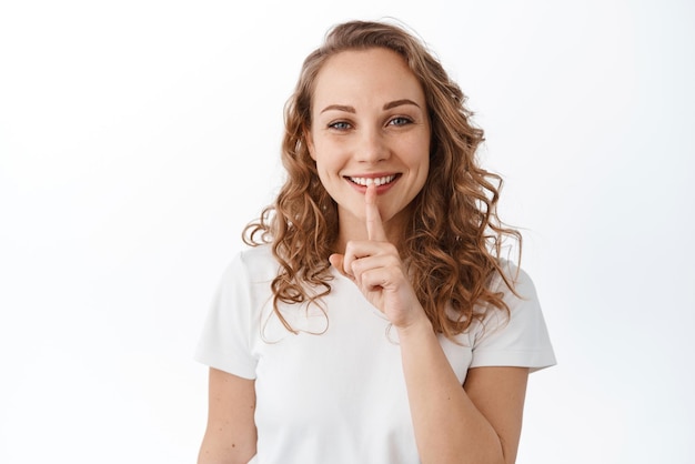 Linda garota sorridente pedindo para ficar quieta fazendo gesto de shh shush pressione o dedo nos lábios e pareça feliz contando segredo em pé sobre fundo branco