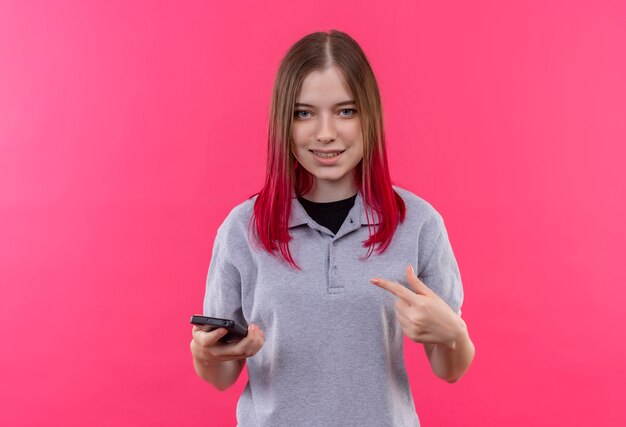 Linda garota sorridente, vestindo uma camiseta cinza, aponta o dedo para o telefone na mão sobre fundo rosa isolado com espaço de cópia