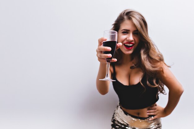 Linda garota sorrindo e levantando a taça de vinho no brinde. Ela está usando saia com lantejoulas, top preto. Look sexy e elegante com barriga nua e decote profundo. Concentre-se em um copo de vinho tinto. Isolado.