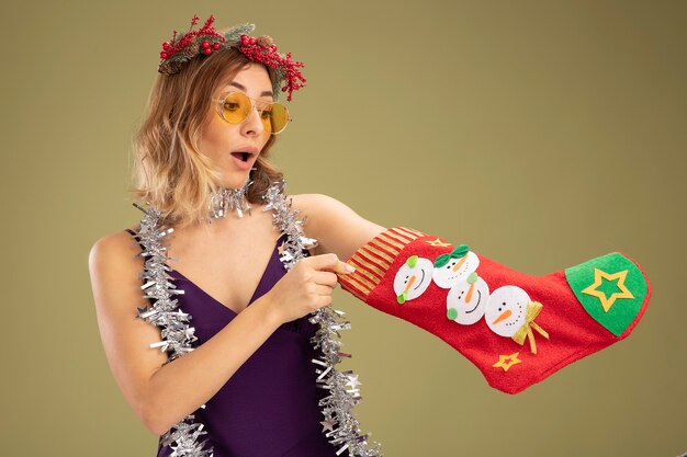 Linda garota surpresa com um vestido roxo com óculos e grinalda com guirlanda no pescoço colocando a mão na meia de natal isolada em fundo verde oliva