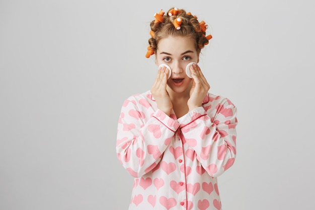 Foto grátis linda garota tirando a maquiagem antes de dormir com almofada de algodão, usando rolinhos de cabelo e pijama