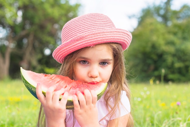 Linda garota usando chapéu-de-rosa comendo fatia de melancia no parque