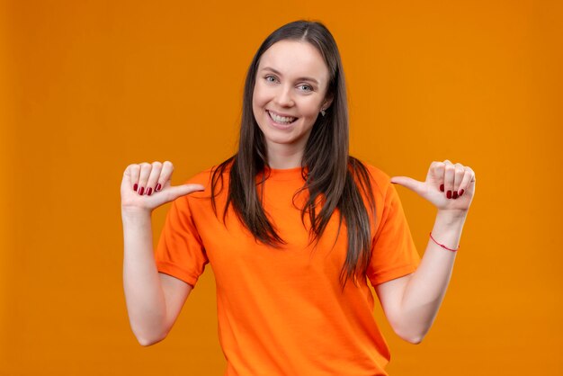Linda garota vestindo uma camiseta laranja apontando para si mesma, satisfeita e orgulhosa, sorrindo em pé sobre um fundo laranja isolado