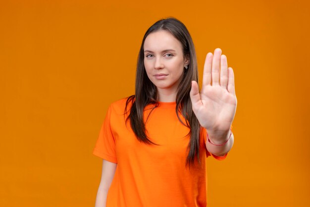Linda garota vestindo uma camiseta laranja em pé com a mão aberta fazendo um gesto de parada em pé sobre um fundo laranja
