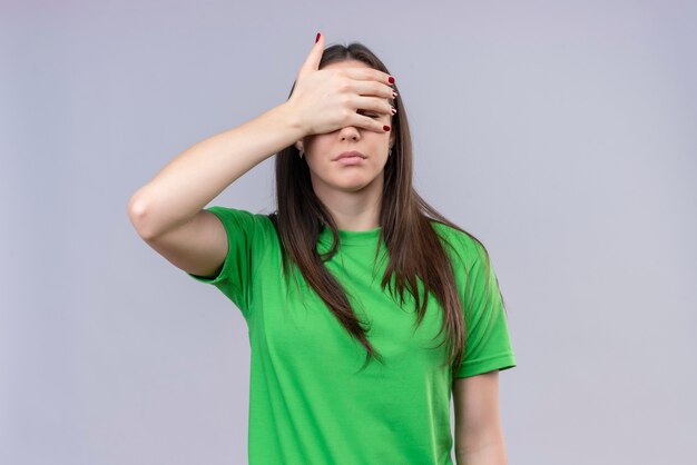 Linda garota vestindo uma camiseta verde cobrindo os olhos com a mão olhando por entre os dedos em pé sobre um fundo branco isolado