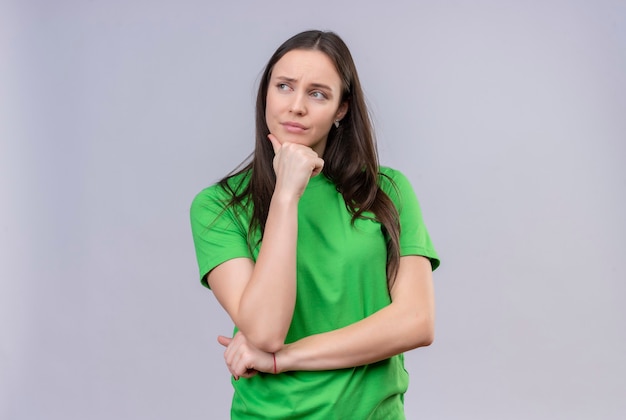 Linda garota vestindo uma camiseta verde em pé com a mão no queixo olhando para o lado com uma expressão pensativa no rosto em pé sobre um fundo branco isolado