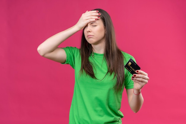 Linda garota vestindo uma camiseta verde segurando um cartão de crédito segurando sua cabeça com a mão por engano chateada com os olhos fechados em pé sobre um fundo rosa isolado