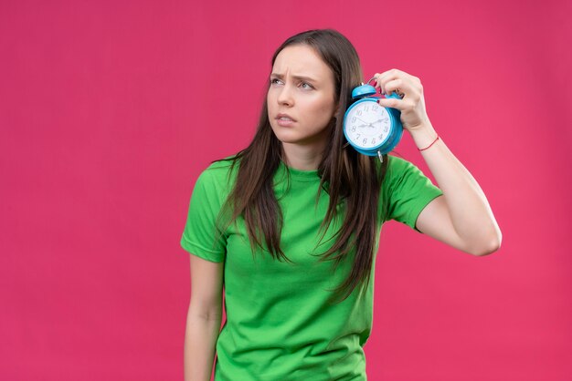 Linda garota vestindo uma camiseta verde segurando um despertador, parecendo confusa em pé sobre um fundo rosa isolado