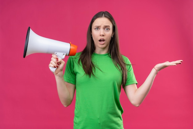 Foto grátis linda garota vestindo uma camiseta verde segurando um megafone, abrindo os braços, parecendo confusa em pé sobre um fundo rosa isolado