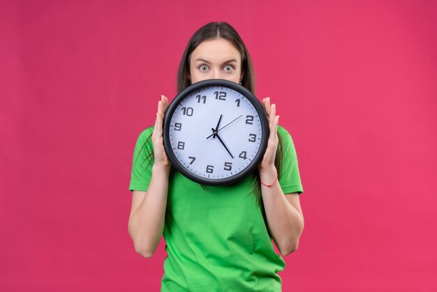 Linda garota vestindo uma camiseta verde segurando um relógio olhando por cima e parecendo surpresa em pé sobre um fundo rosa isolado